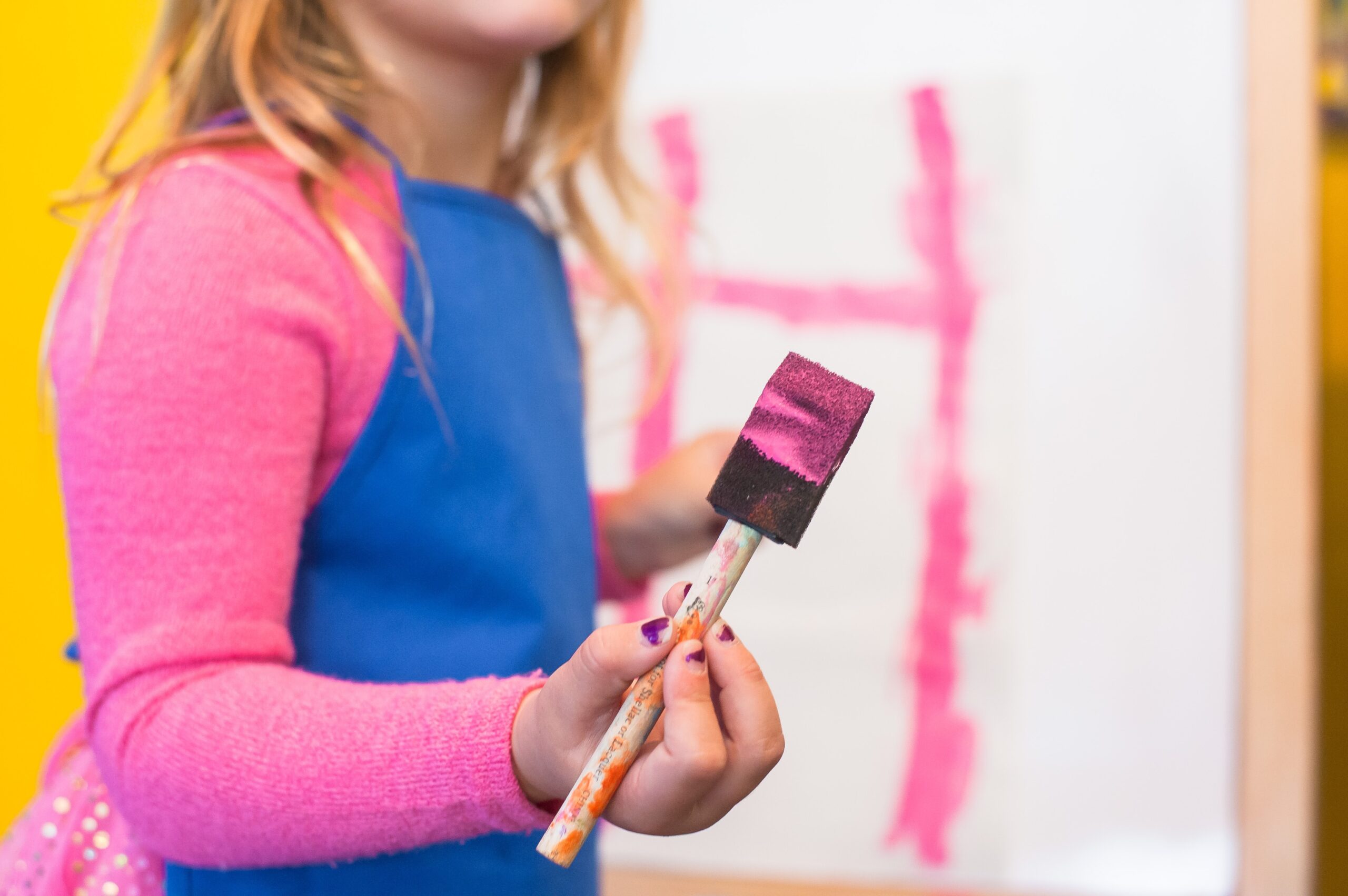 girl in smock holding paintbrush with pink paint on it